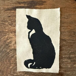Puede incluir: Silueta negra de un gato sentado sobre un fondo de tela beige.