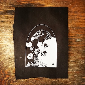 May include: A black and white patch featuring a stylised image of a vulture's head within an archway, surrounded by flowers. The patch is made of fabric and has a rough texture.