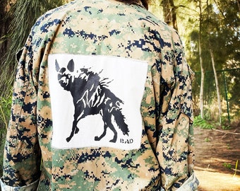 May include: A camouflage jacket with a black and white patch of a hyena on the back. The patch has the word "BAD" printed below the hyena.