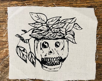 May include: A black and white linocut print on a square piece of fabric. The print depicts a skull with a plant growing out of the top. The plant has many leaves. The fabric has a raw edge.