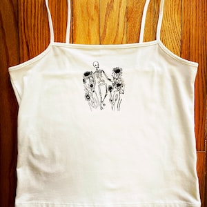 May include: White spaghetti strap tank top with a black line drawing of a skeleton standing in a field of sunflowers.