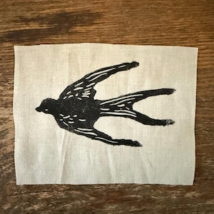 May include: A black bird illustration in flight is printed on a square of natural-colored fabric. The bird's wings are outstretched, and its body is detailed with white lines and dots. The fabric has a slightly wrinkled texture.