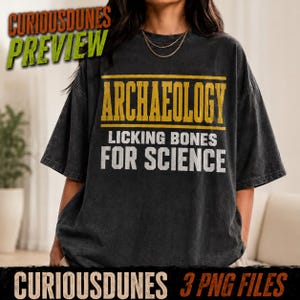 以下が含まれることがあります： 黄色い文字で「ARCHAEOLOGY」、白い文字で「LICKING BONES FOR SCIENCE」とプリントされたチャコールグレーの半袖Tシャツ。クルーネックでゆったりとしたフィット感があり、全体的に色あせたようなヴィンテージ感のある生地の質感が特徴です。