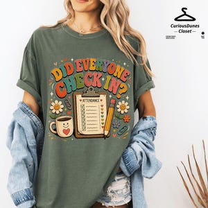 May include: An olive green t-shirt featuring a graphic with the text DID EVERYONE CHECK IN? above an attendance clipboard, a smiling coffee mug, a pencil, paper clips, and flowers in shades of orange, yellow, blue, and red on the front.