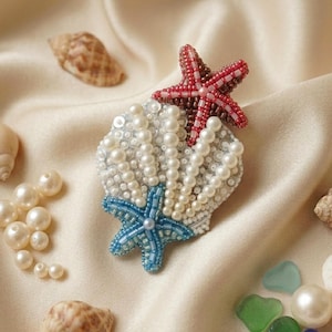 Op de afbeelding: Een handgemaakte broche in de vorm van een schelp, versierd met witte parelkralen en lovertjes. Een rode kraalster is aan de bovenkant bevestigd en een blauwe kraalster aan de onderkant, wat zorgt voor een gestructureerd, veelkleurig accessoire.
