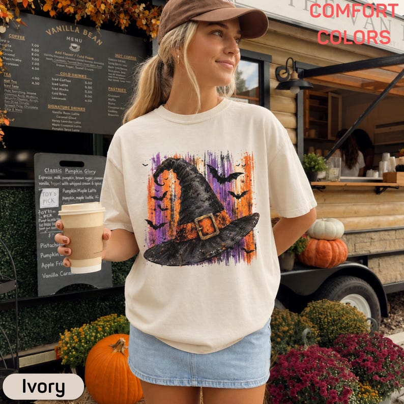 May include: An ivory short-sleeve t-shirt featuring a graphic print of a black witch hat with an orange buckle, surrounded by purple and orange brushstrokes and black bats. The text Comfort Colors is visible in the upper right corner.
