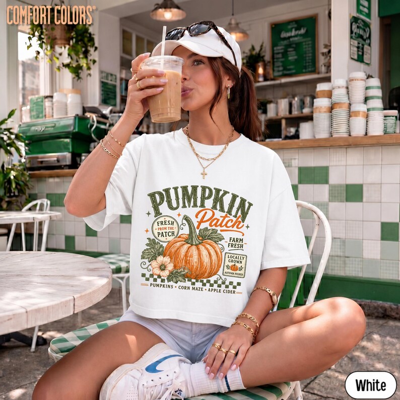 Op de afbeelding: Een wit T-shirt met korte mouwen en een grafische print van een oranje pompoen met de tekst Pumpkin Patch, Fresh From The Patch, Farm Fresh, Pumpkins, Corn Maze en Apple Cider. Het T-shirt heeft een ronde halslijn.