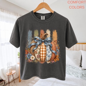 Könnte beinhalten: Ein anthrazitfarbenes Kurzarm-T-Shirt mit einer Grafik eines Kürbisses mit verschiedenen Mustern wie Leopardenmuster, Karos und Blumen, gebunden mit einer blauen Schleife. Der Text COMFORT COLORS steht in der oberen rechten Ecke.