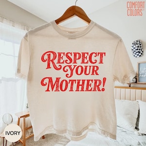 May include: An ivory short-sleeve t-shirt featuring the red text "RESPECT YOUR MOTHER!" printed in a bold, stylized font across the chest. The garment has a crew neckline and is displayed on a wooden hanger against a neutral indoor setting.