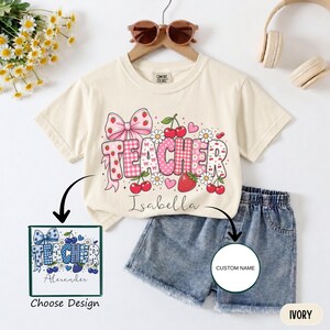 May include: An ivory short-sleeve t-shirt featuring a graphic with the word TEACHER in pink and white patterns, decorated with strawberries, cherries, daisies, and a large pink bow. The name Isabella is printed below, with a blue design option shown.