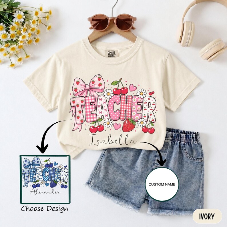 May include: An ivory short-sleeve t-shirt featuring a graphic with the word TEACHER in pink and white patterns, decorated with strawberries, cherries, daisies, and a large pink bow. The name Isabella is printed below, with a blue design option shown.