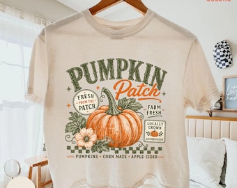 Op de afbeelding: Een ivoorkleurig t-shirt met korte mouwen en een grafische print van een grote oranje pompoen met groene bladeren. De tekst luidt PUMPKIN Patch, FRESH FROM THE PATCH, FARM FRESH, LOCALLY GROWN en PUMPKINS CORN MAZE APPLE CIDER in diverse lettertypes.