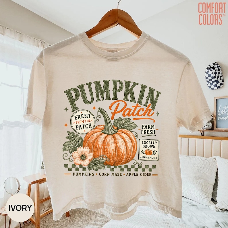 Op de afbeelding: Een ivoorkleurig t-shirt met korte mouwen en een grafische print van een grote oranje pompoen met groene bladeren. De tekst luidt PUMPKIN Patch, FRESH FROM THE PATCH, FARM FRESH, LOCALLY GROWN en PUMPKINS CORN MAZE APPLE CIDER in diverse lettertypes.
