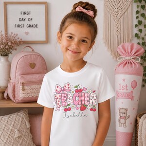 May include: A white short-sleeved t-shirt featuring a graphic print with the word TEACHER in pink and white patterned letters, decorated with cherries, strawberries, daisies, and a bow. The name Isabella is printed in cursive below the graphic.