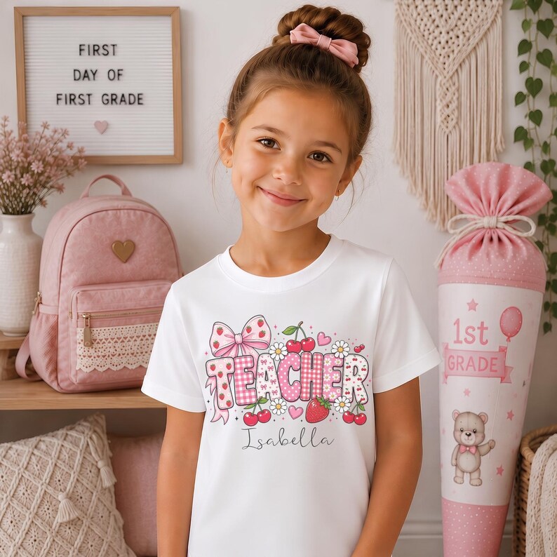 May include: A white short-sleeved t-shirt featuring a graphic print with the word TEACHER in pink and white patterned letters, decorated with cherries, strawberries, daisies, and a bow. The name Isabella is printed in cursive below the graphic.