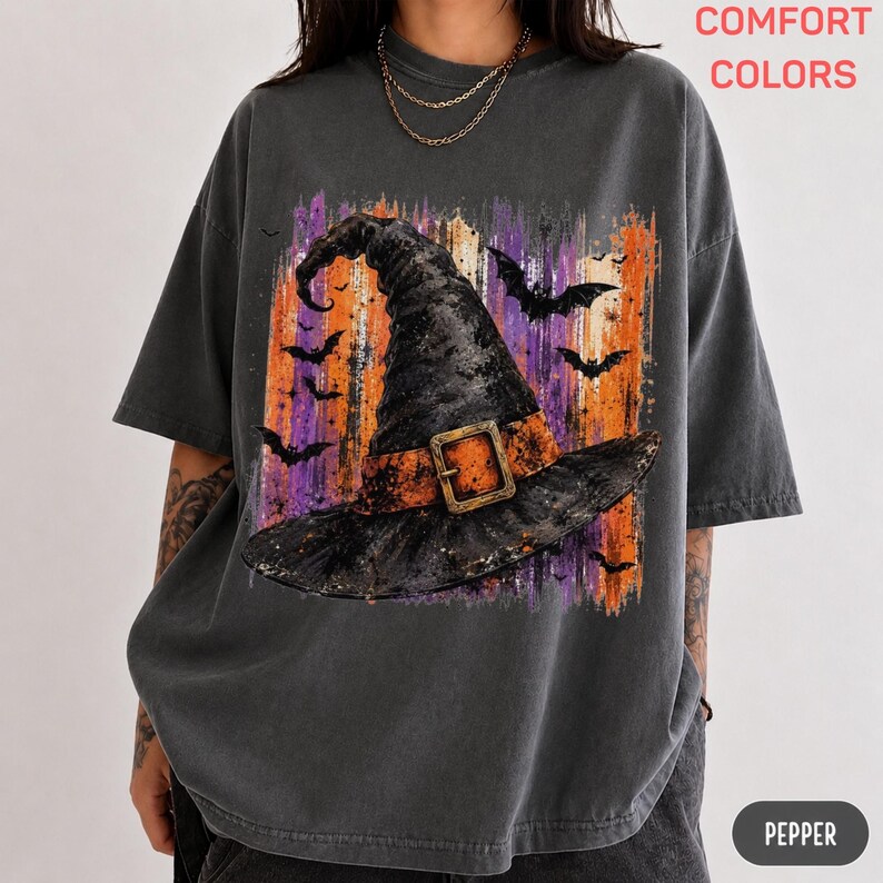May include: A charcoal gray short-sleeve t-shirt featuring a graphic of a black witch hat with an orange band and buckle, set against a background of vertical orange and purple brushstrokes with black bat silhouettes and the text COMFORT COLORS.