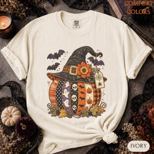 May include: An ivory t-shirt featuring a graphic of a pumpkin wearing a witch hat. The pumpkin has vertical panels with ghosts, skulls, and jack-o'-lanterns. The hat is decorated with a sunflower, bats, and a spider web tag. Comfort Colors label.