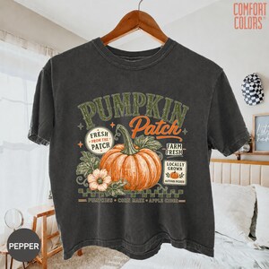 Op de afbeelding: Een antracietgrijs T-shirt met korte mouwen en een grafische print van een grote oranje pompoen met groene bladeren. De tekst luidt PUMPKIN Patch, FRESH FROM THE PATCH, FARM FRESH, LOCALLY GROWN, PUMPKINS, CORN MAZE en APPLE CIDER.