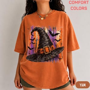 May include: An orange short-sleeve t-shirt featuring a graphic print of a black witch hat with a buckle, surrounded by silhouettes of flying bats against a background of vertical purple and orange brushstrokes. The shirt has a crew neckline.