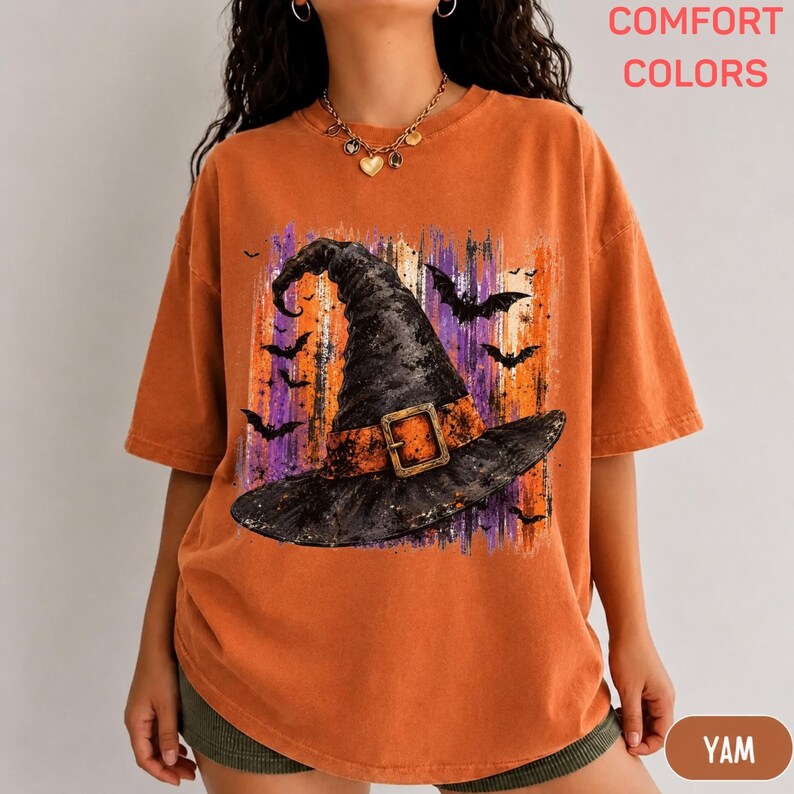 May include: An orange short-sleeve t-shirt featuring a graphic print of a black witch hat with a buckle, surrounded by silhouettes of flying bats against a background of vertical purple and orange brushstrokes. The shirt has a crew neckline.
