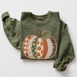 May include: An olive green crewneck sweatshirt featuring a large, central pumpkin graphic. The pumpkin is composed of vertical fabric panels with various patterns, including orange gingham, floral prints, and herringbone, with a green leaf.