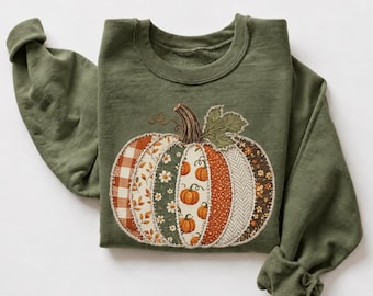 Op de afbeelding: Een olijfgroene sweater met ronde hals en een grote, centrale pompoenafbeelding. De pompoen is samengesteld uit verticale stoffen panelen met diverse patronen, waaronder oranje gingham, bloemenprints en visgraat, met een groen blad.