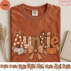 May include: A rust-colored crew neck t-shirt with a graphic print featuring the word Auntie in large letters with various patterns including polka dots, leopard print, and floral designs. The graphic includes two pumpkins and a large bow.