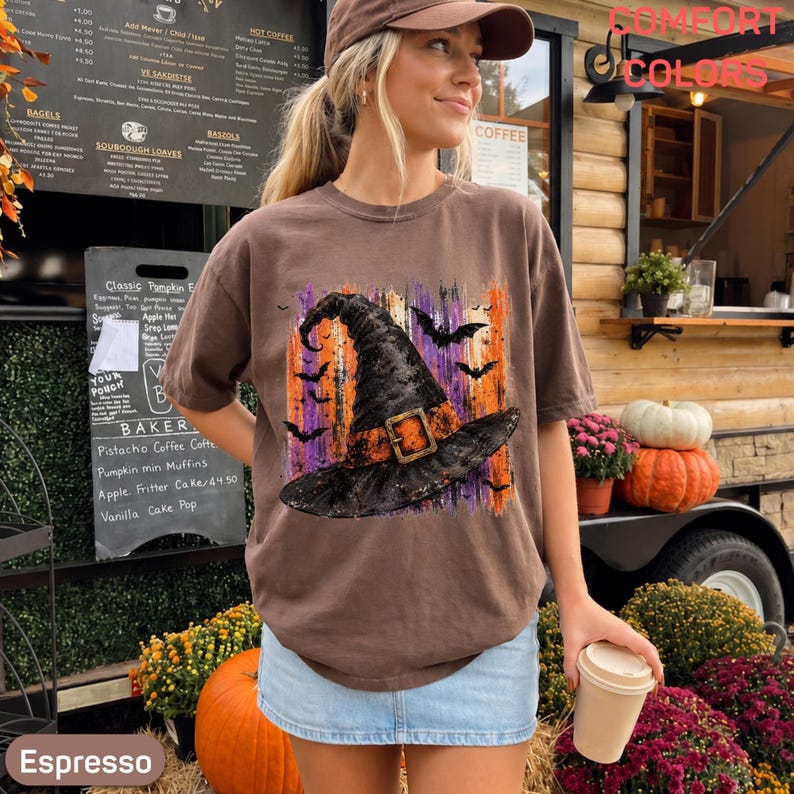 May include: A brown short-sleeve t-shirt featuring a graphic print of a black witch hat with an orange band and buckle, surrounded by black bats and vertical orange and purple brushstroke accents on the front of the garment.