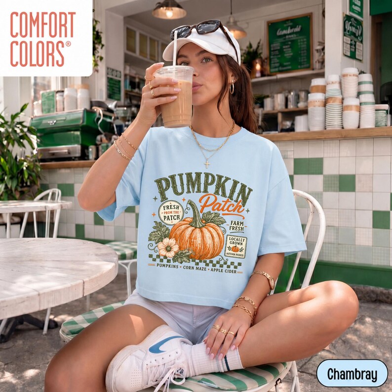 Op de afbeelding: Een lichtblauw t-shirt met een grafische print van een grote oranje pompoen met de tekst Pumpkin Patch, Fresh From The Patch, Farm Fresh en Locally Grown. Het shirt bevat een dambordpatroon en kleine illustraties van pompoenen.