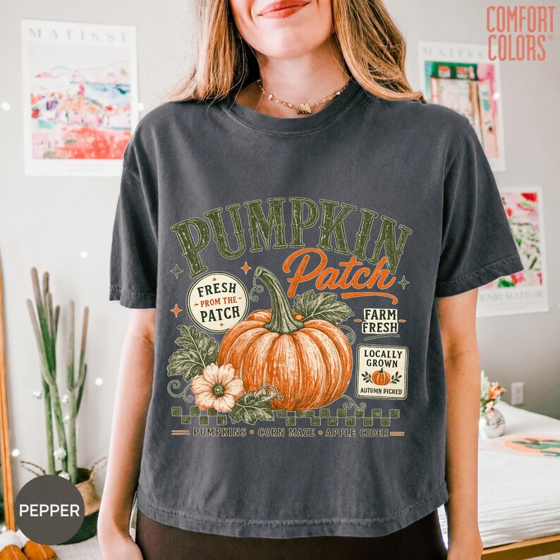 Op de afbeelding: Een antracietgrijs T-shirt met korte mouwen en een grafische print van een grote oranje pompoen met groene bladeren. De tekst luidt PUMPKIN PATCH, FRESH FROM THE PATCH, FARM FRESH, LOCALLY GROWN, PUMPKINS, CORN MAZE en APPLE CIDER.