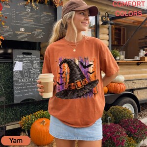 May include: A rust-colored short-sleeve t-shirt featuring a graphic print of a black witch hat with a gold buckle, surrounded by purple and orange vertical brushstrokes and black bat silhouettes. The shirt has a crew neckline and a loose fit.