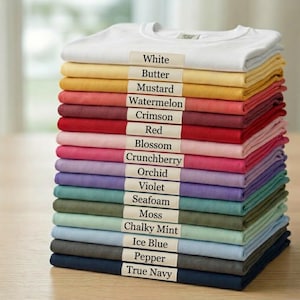 May include: A vertical stack of folded t-shirts in various colors, each featuring a small rectangular label with the color name printed in black text. The colors include white, butter, mustard, watermelon, crimson, red, blossom, crunchberry, orchid, violet, seafoam, moss, chalky mint, ice blue, pepper, and true navy.