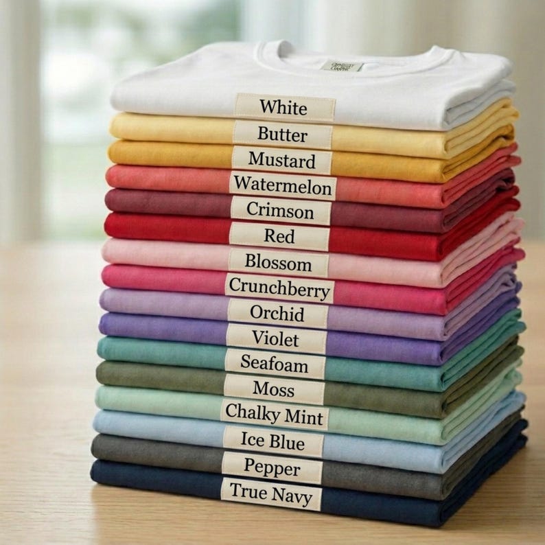May include: A vertical stack of folded t-shirts in various colors, each featuring a small rectangular label with the color name printed in black text. The colors include white, butter, mustard, watermelon, crimson, red, blossom, crunchberry, orchid, violet, seafoam, moss, chalky mint, ice blue, pepper, and true navy.