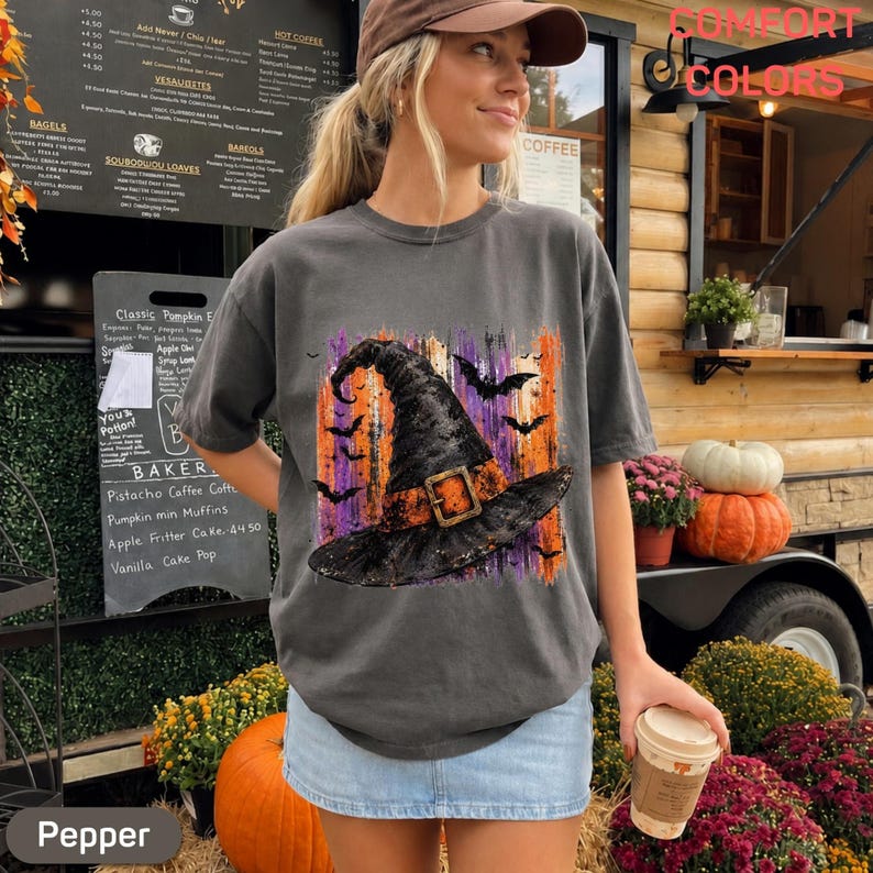 May include: A charcoal gray t-shirt featuring a graphic of a black witch hat with an orange band and buckle, surrounded by silhouettes of flying bats against a vertical brushstroke background of purple, orange, and black colors.