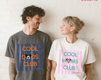 Op de afbeelding: Een antracietgrijs T-shirt met blauwe verticale strepen, oranje tekst COOL DADS CLUB, Mickey Mouse-icoon en de naam Stephen, naast een wit T-shirt met roze verticale strepen, blauwgroene tekst COOL MOMS CLUB, Minnie Mouse-icoon en de naam Jennifer.