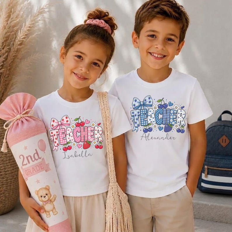 May include: Two white t-shirts featuring the word TEACHER in patterned block letters, decorated with cherries, strawberries, and bows. One shirt includes the name Isabella, the other Alexander. A pink cone-shaped gift bag labeled 2nd Grade is held.