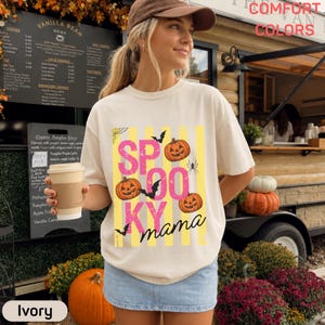 Puede incluir: Una camiseta de color marfil con un gráfico que muestra el texto SPOOKY mama en letras rosas y negras. El diseño incluye tres calabazas naranjas, murciélagos negros y una araña, sobre un fondo de rayas verticales de color amarillo.