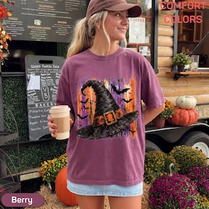 May include: A berry-colored short-sleeve t-shirt featuring a graphic print of a black witch hat with an orange buckle, surrounded by silhouettes of flying bats against a vertical orange and purple brushstroke background.
