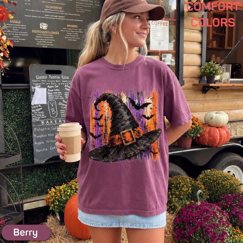 May include: A berry-colored short-sleeve t-shirt featuring a graphic print of a black witch hat with an orange buckle, surrounded by silhouettes of flying bats against a vertical orange and purple brushstroke background.