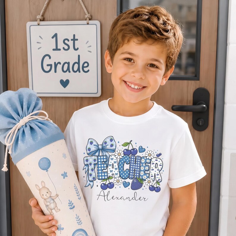 May include: A white short-sleeved t-shirt featuring a blue patterned graphic with the word TEACHER, cherries, strawberries, and a bow, with the name Alexander printed below. Beside it is a light blue and cream cone-shaped gift bag.