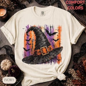 May include: An ivory t-shirt featuring a graphic of a black witch hat with an orange band and buckle. The design is set against a background of vertical orange and purple brushstrokes with black bat silhouettes. The inner label reads Comfort Colors.
