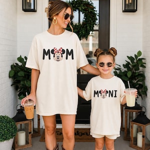 May include: Two cream-colored short-sleeve t-shirts featuring black text and a Minnie Mouse graphic. The larger shirt displays the word MOM with a Minnie Mouse icon replacing the letter O, while the smaller shirt displays the word MINI with a Minnie Mouse icon.