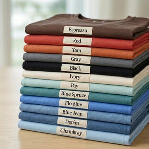 May include: A stack of folded t-shirts in various colors including espresso, red, yam, gray, black, ivory, bay, blue spruce, flo blue, blue jean, denim, and chambray. Each shirt features a small rectangular label with the color name printed.