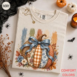 May include: An ivory t-shirt featuring a graphic of a pumpkin with segments in leopard print, orange gingham, floral, and polka dot patterns. A large blue ribbon is tied around the pumpkin, surrounded by autumn leaves and flowers.