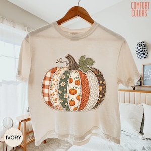 May include: An ivory short-sleeve t-shirt featuring a large graphic of a pumpkin composed of vertical panels with various patterns including plaid, floral, and herringbone prints in shades of orange, green, brown, and white on the front.