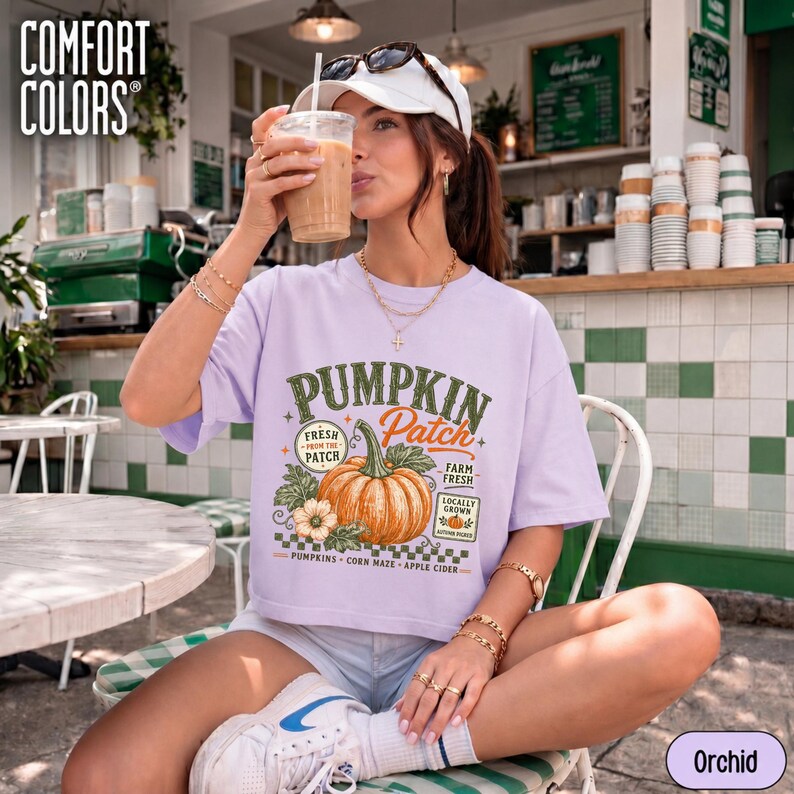 Op de afbeelding: Een lichtpaars t-shirt met een afbeelding van een grote oranje pompoen met groene bladeren en ranken. De tekst luidt PUMPKIN Patch, FRESH FROM THE PATCH, FARM FRESH en PUMPKINS CORN MAZE APPLE CIDER. Het t-shirt wordt gedragen met een witte pet.