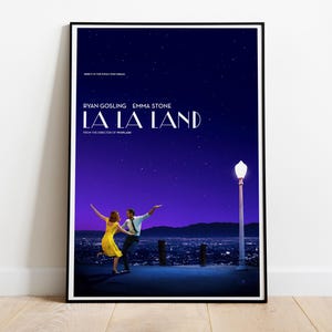 May include: A framed film poster for La La Land featuring a dark blue starry night sky over a city landscape. Two figures are dancing on a platform near a street lamp. The text reads: Here's to the fools who dream, Ryan Gosling, Emma Stone, La La Land.