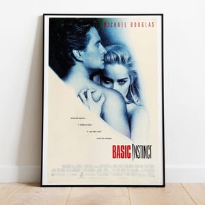May include: A framed movie poster for Basic Instinct featuring a blue-toned, high-contrast photograph of two people embracing. The title Basic Instinct is printed in bold red and black text at the bottom, with Michael Douglas at the top.