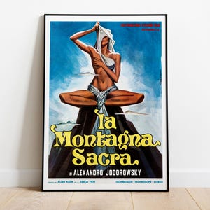 May include: A framed movie poster for La Montagna Sacra by Alexandro Jodorowsky. The artwork features a figure in a meditative pose atop a dark mountain against a blue sky, draped in white fabric. The title is in large yellow lettering at the bottom.