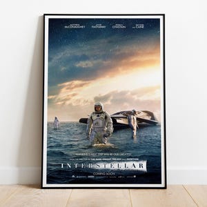 Op de afbeelding: Een ingelijste filmposter voor Interstellar met een astronaut die in ondiep water staat met een ruimteschip in de buurt. De lucht gaat over van een donkere sterrennacht naar een heldere horizon. De tekst bevat de namen van de acteurs.